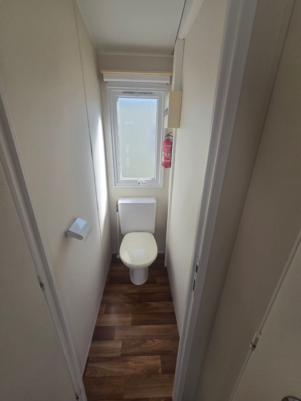 Cabine de toilettes étroite avec cuvette blanche, fenêtre dépolie et sol à motif bois.