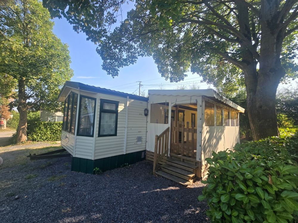 Maison mobile blanche avec marches en bois et porche dans une cour arborée, entourée d'arbres et de gravier.