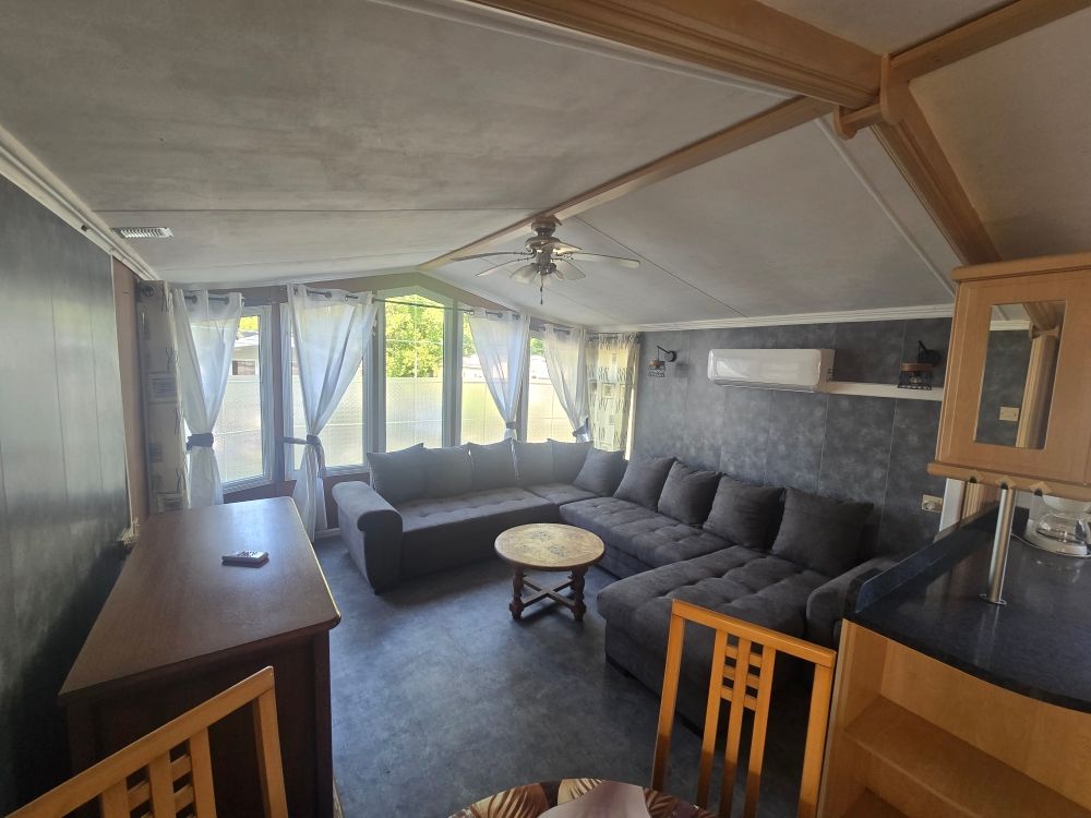 Véranda confortable avec canapé d'angle gris, table ronde et grandes fenêtres avec voilages.
