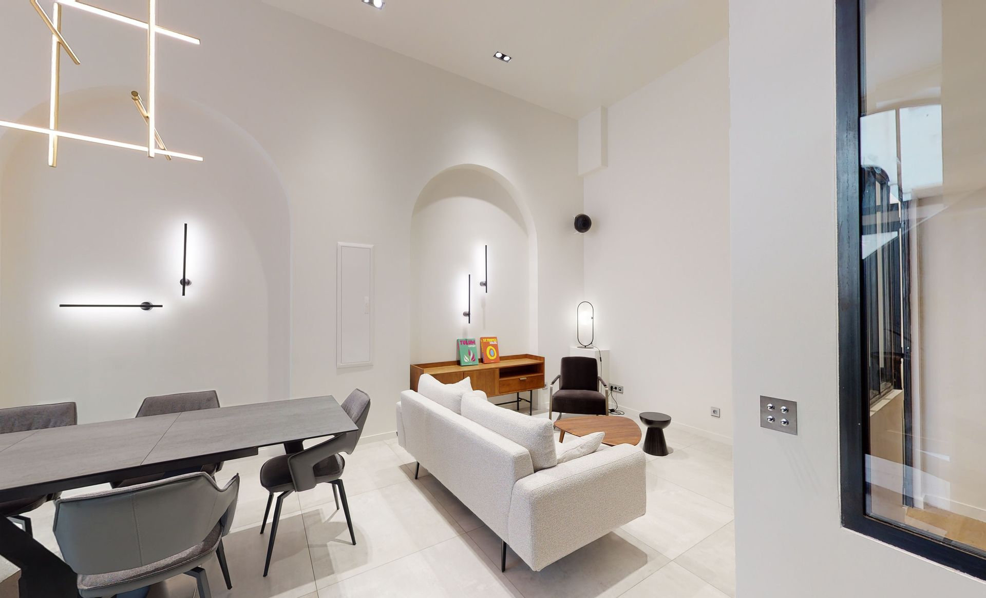 Salon rénové avec murs blancs et canapé beige ainsi qu'une table à manger avec des chaises anthracites