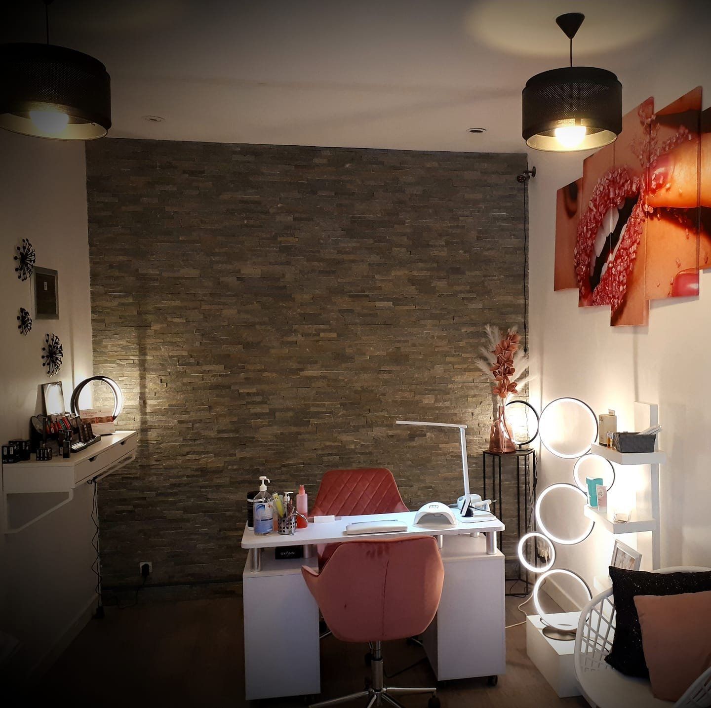 Intérieur de salon de manucure avec un bureau, une chaise rose, un mur en pierre en arrière-plan et un éclairage décoratif.