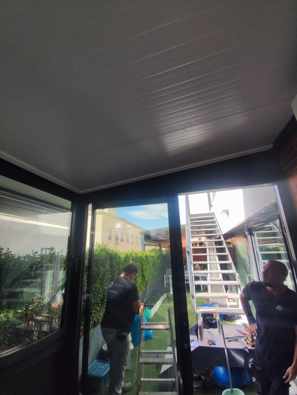 Dos personas en un patio cubierto, trabajando cerca de una escalera y cubos fuera de una puerta de cristal.