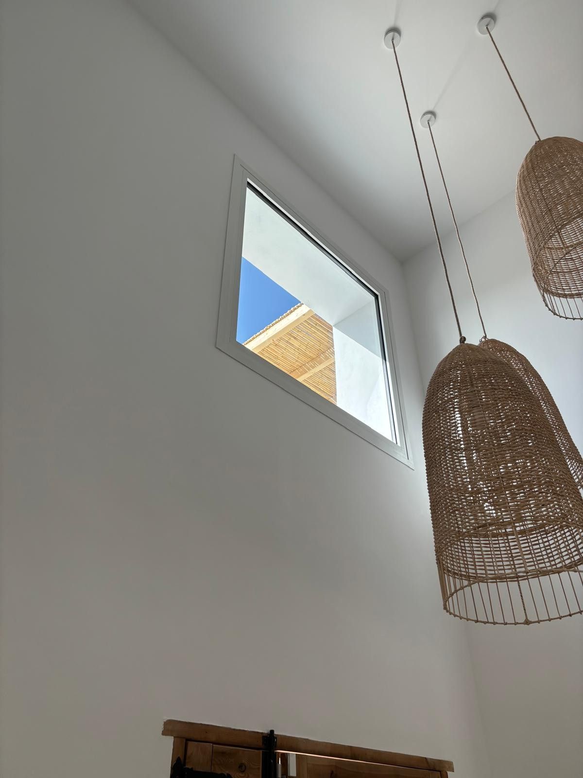 Pared blanca alta con una pequeña ventana, cielo azul afuera y lámparas colgantes tejidas en el interior.