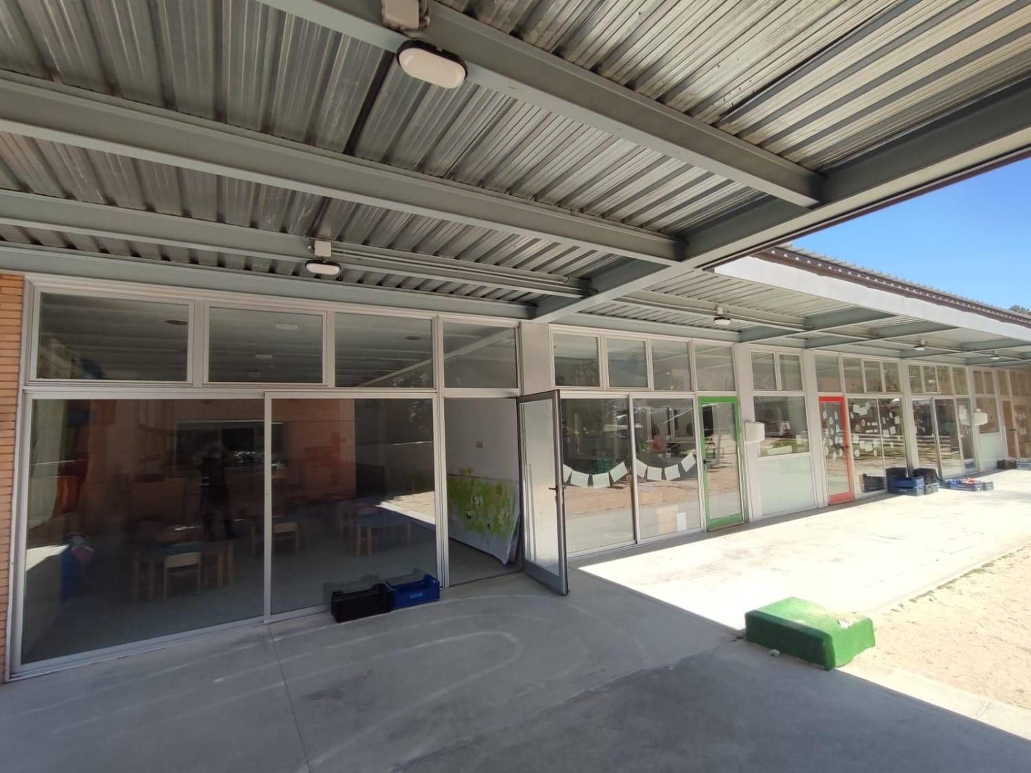 Patio exterior cubierto con escaparates de cristal, suelo de hormigón y un banco verde bañado por la luz del sol.