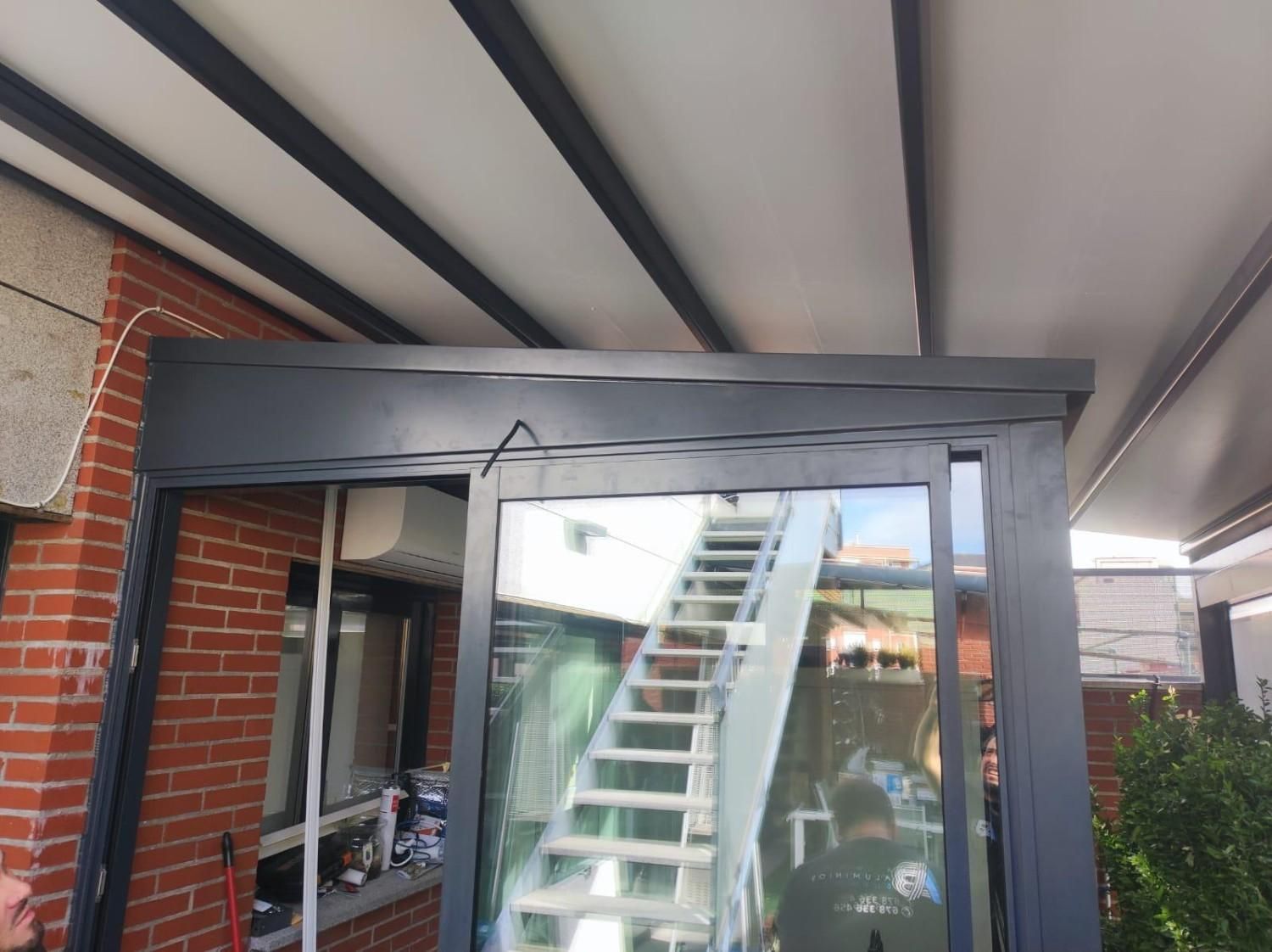 Patio acristalado con una escalera blanca en su interior, visible desde un techo cubierto junto a una pared de ladrillo.