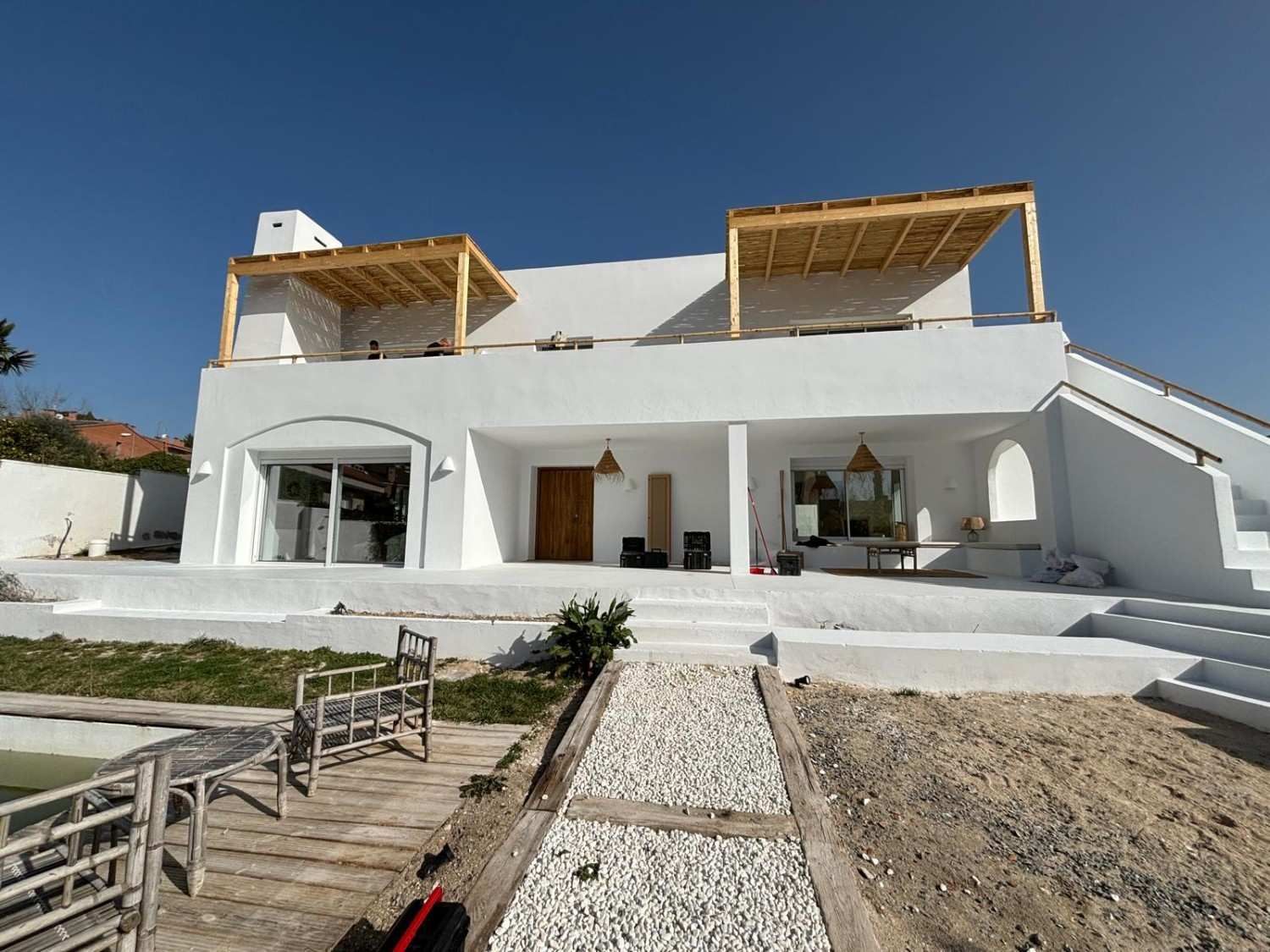 Casa moderna blanca con terrazas de madera abiertas en el techo y un camino de entrada bajo un cielo azul claro.