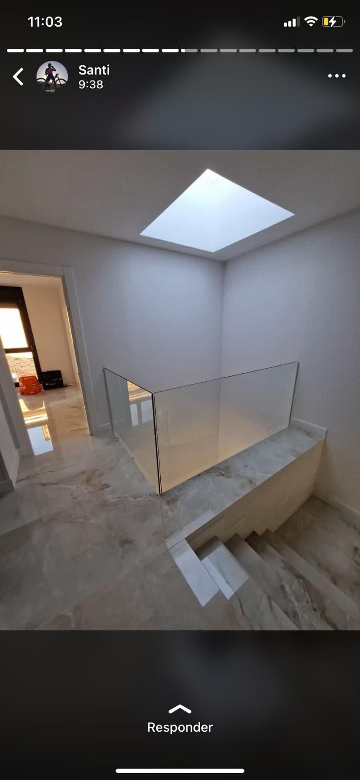 Escalera moderna con paredes blancas, tragaluz, barandilla de cristal y suelo de baldosas en un interior luminoso.
