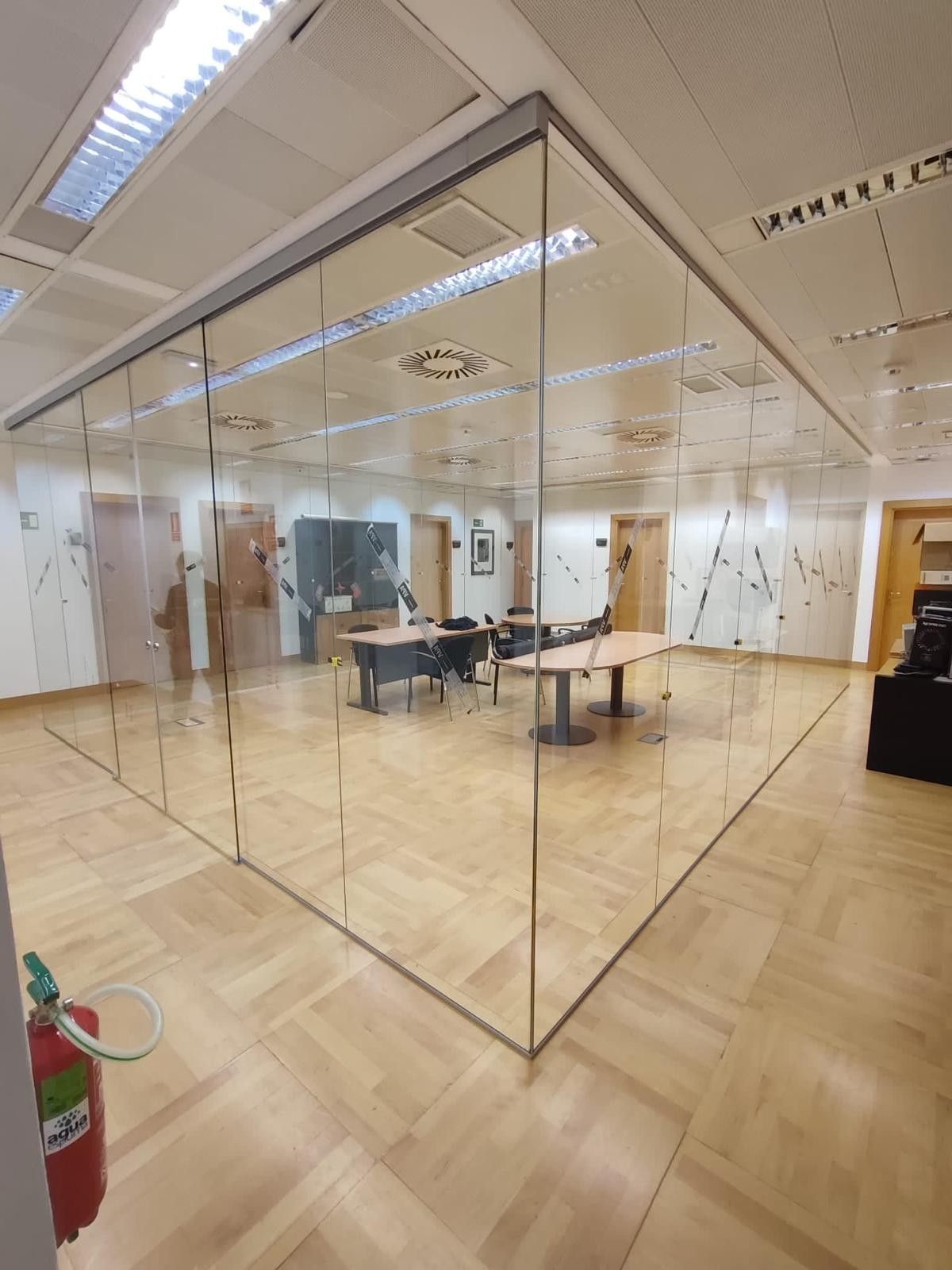 Sala de reuniones de oficina con paredes de cristal, escritorios y sillas sobre un suelo de madera