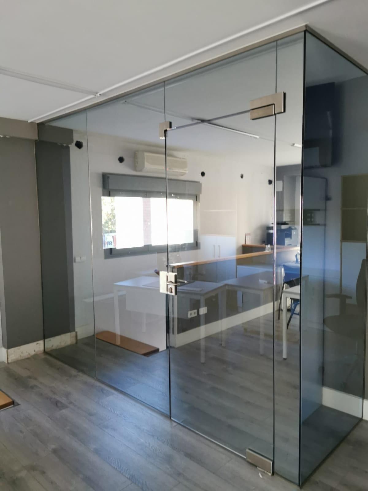 Sala de reuniones de oficina con paredes de cristal, suelo gris, paredes blancas y un escritorio cerca de una ventana.