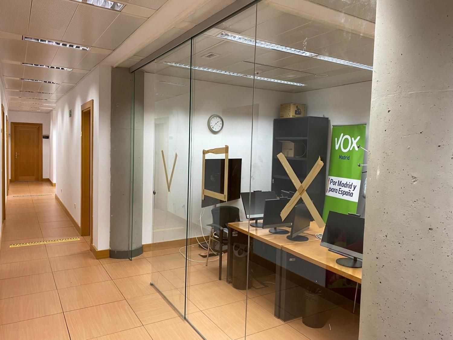 Pasillo de oficina con paredes de cristal, escritorios, monitores de ordenador y un póster verde