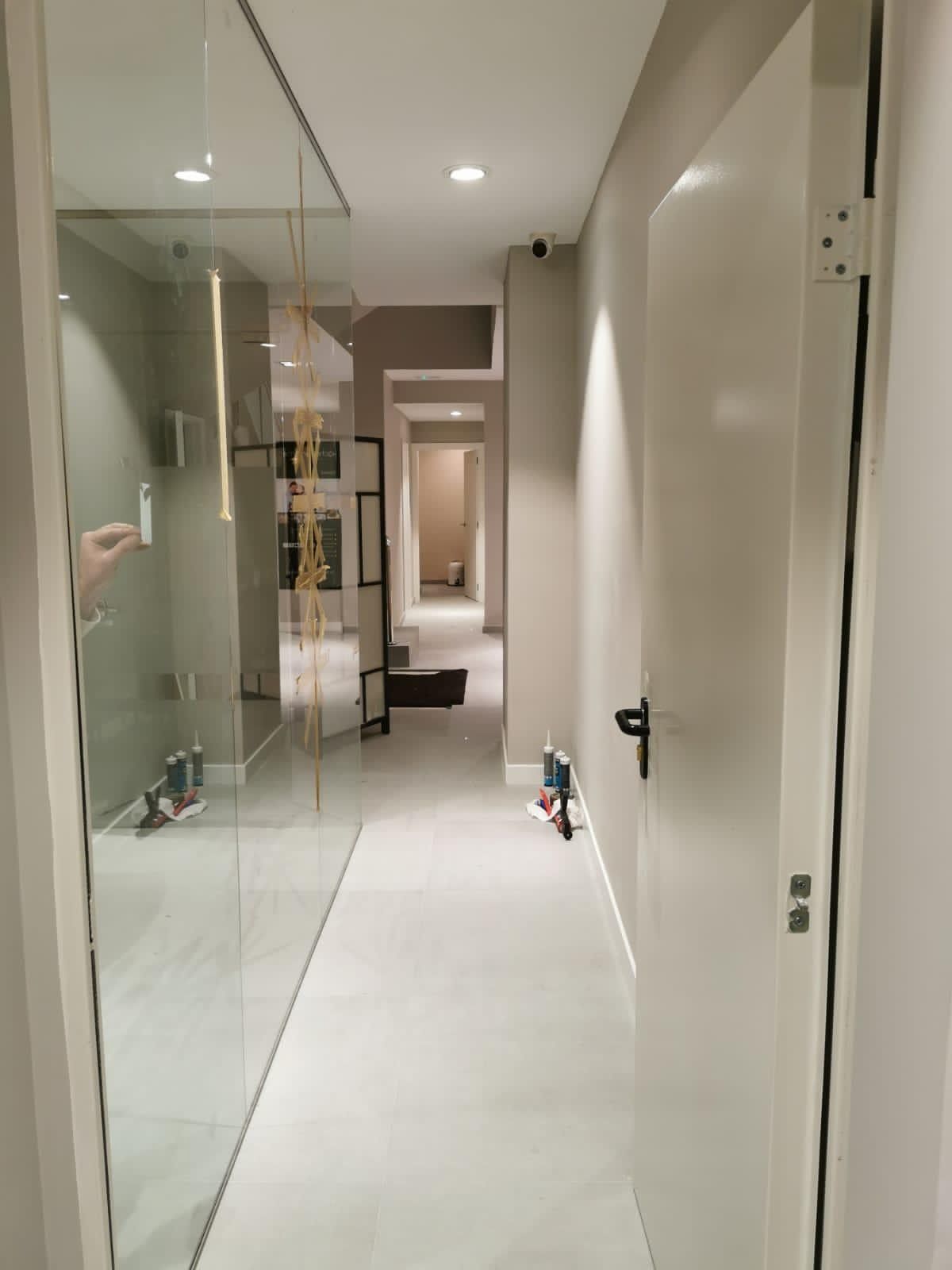 Pasillo moderno y estrecho con pared de cristal, puertas blancas y suelo de baldosas que conduce a una habitación.