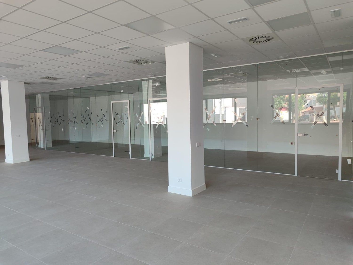 Oficina moderna vacía con paredes de cristal, suelo de baldosas grises y columnas blancas en el techo.