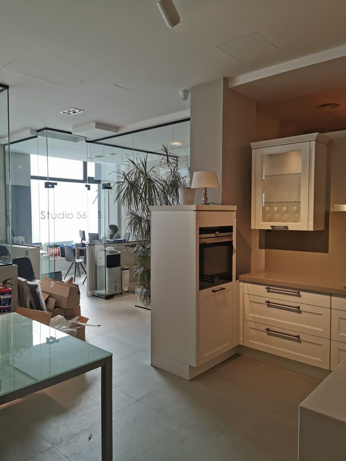 Cocina y espacio de oficina modernos y luminosos con armarios blancos, mesa de cristal y grandes ventanales.