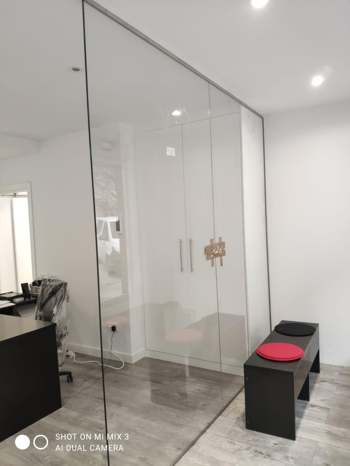 Vestíbulo de oficina moderno con puertas de cristal, paredes blancas, suelo gris y un banco negro con cojines rojos.