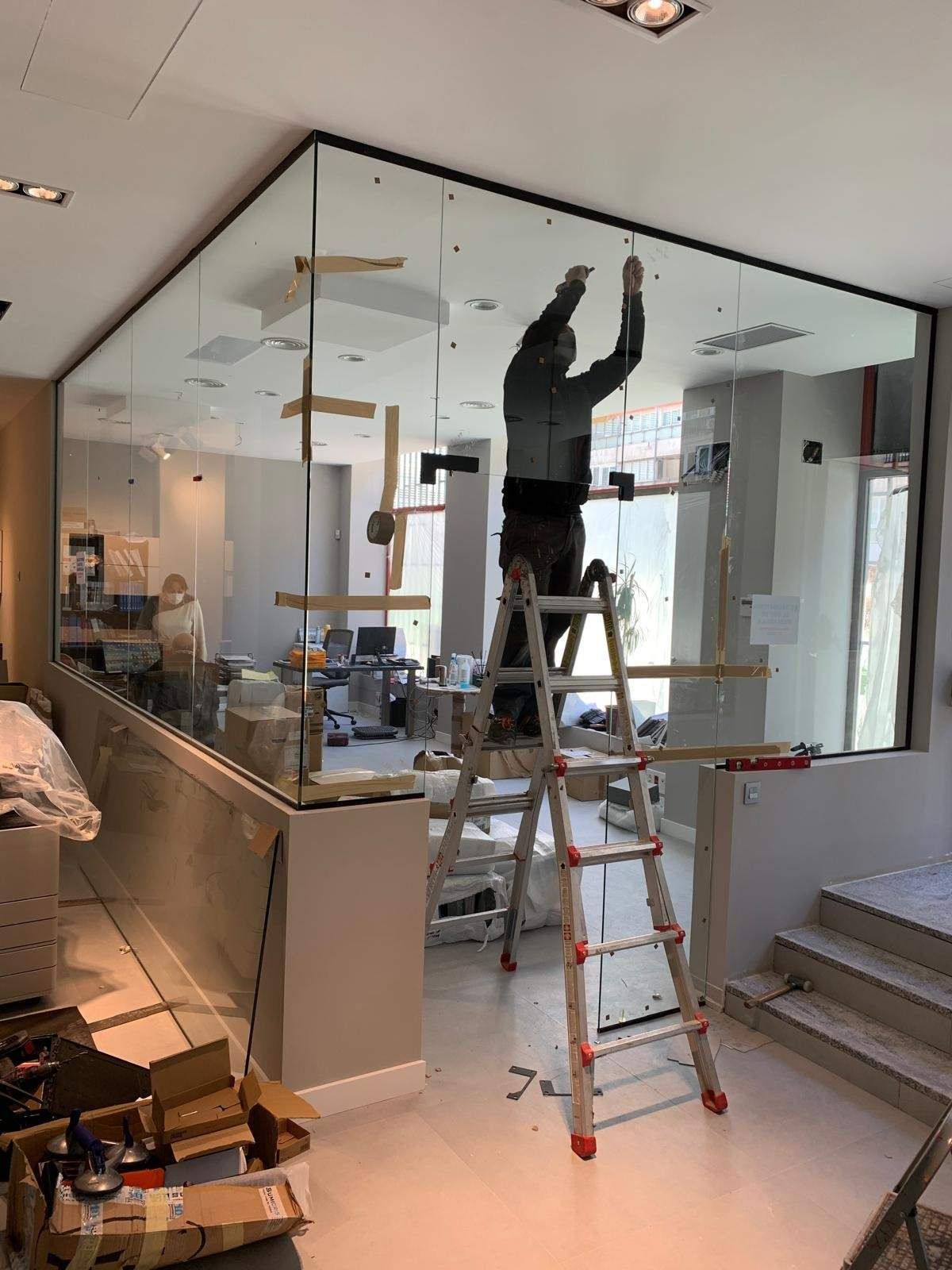 Un trabajador subido a una escalera instala paneles de vidrio en una oficina luminosa.