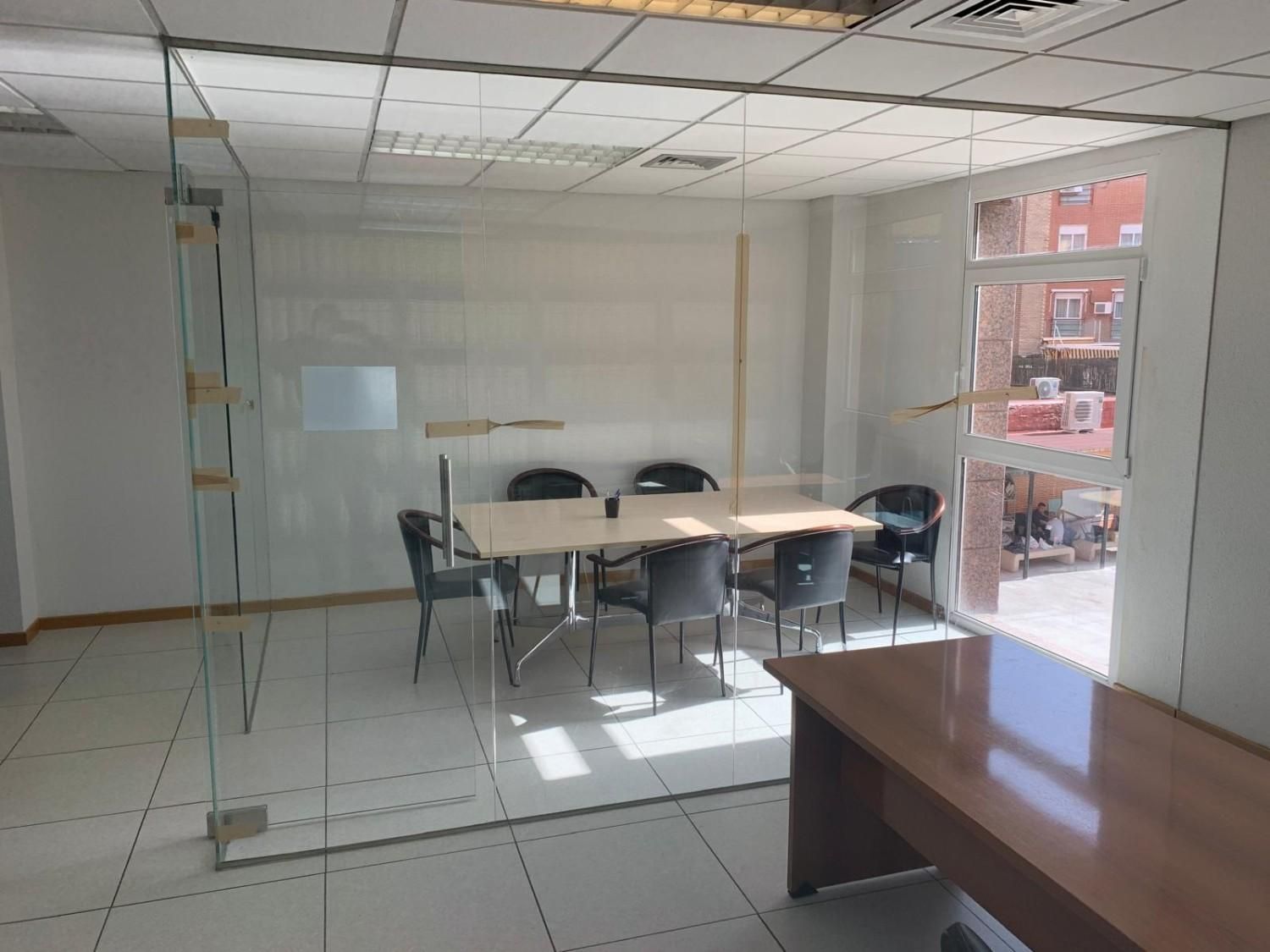 Oficina vacía con paredes de cristal, mesa de conferencias, sillas y un escritorio de madera en primer plano.