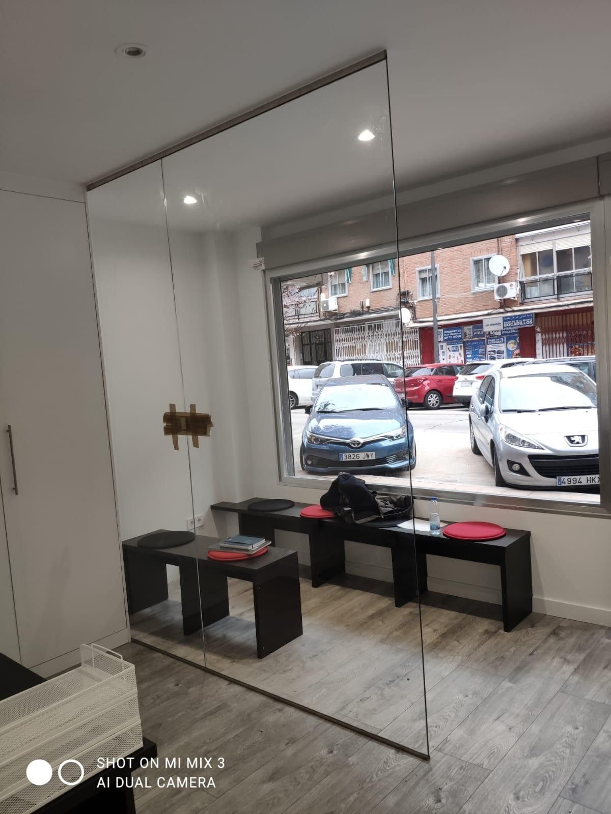 Local comercial vacío con un gran espejo, bancos negros y vista a la calle a través de la ventana frontal.