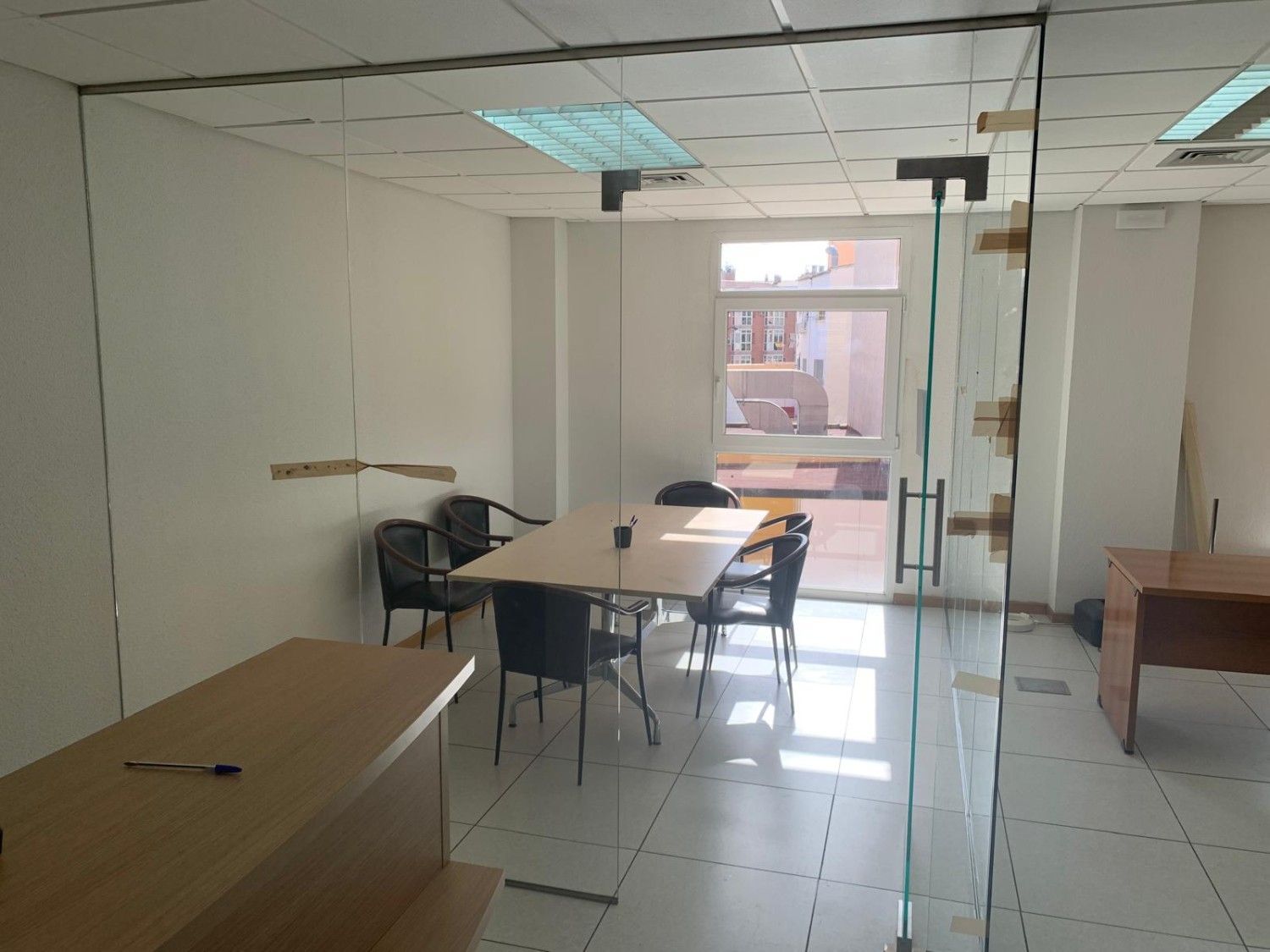 Oficina vacía con escritorios, sillas y una puerta de cristal que da a un pasillo.