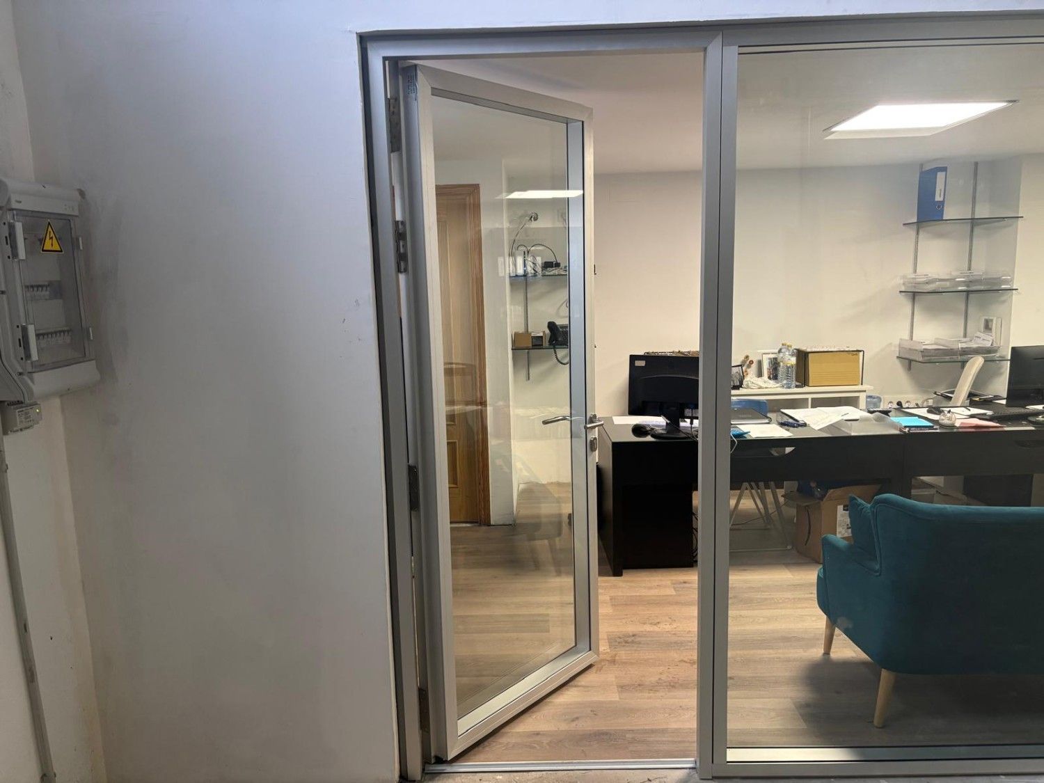 Interior de oficina visto a través de una puerta de cristal abierta, con escritorio, ordenador, estanterías y silla azul.