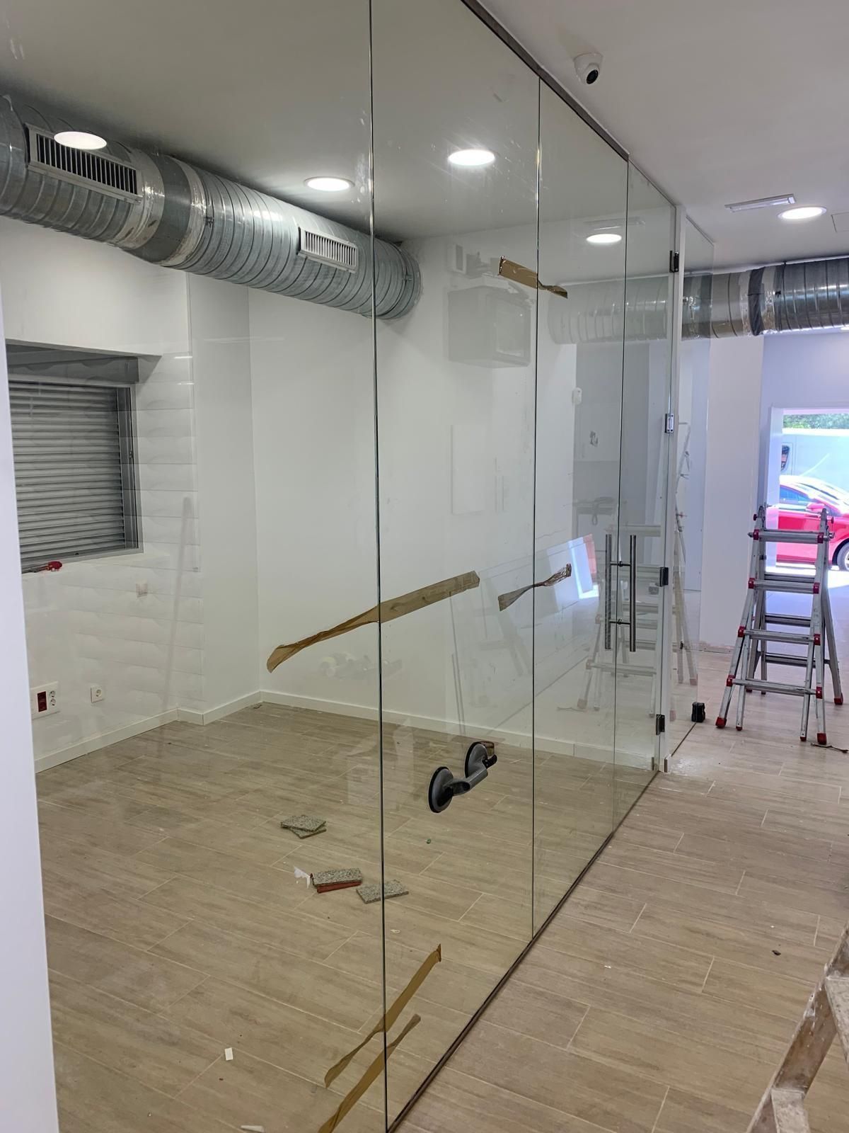 Habitación en construcción con tabiques de vidrio, conductos de ventilación expuestos, escalera y herramientas en el suelo.