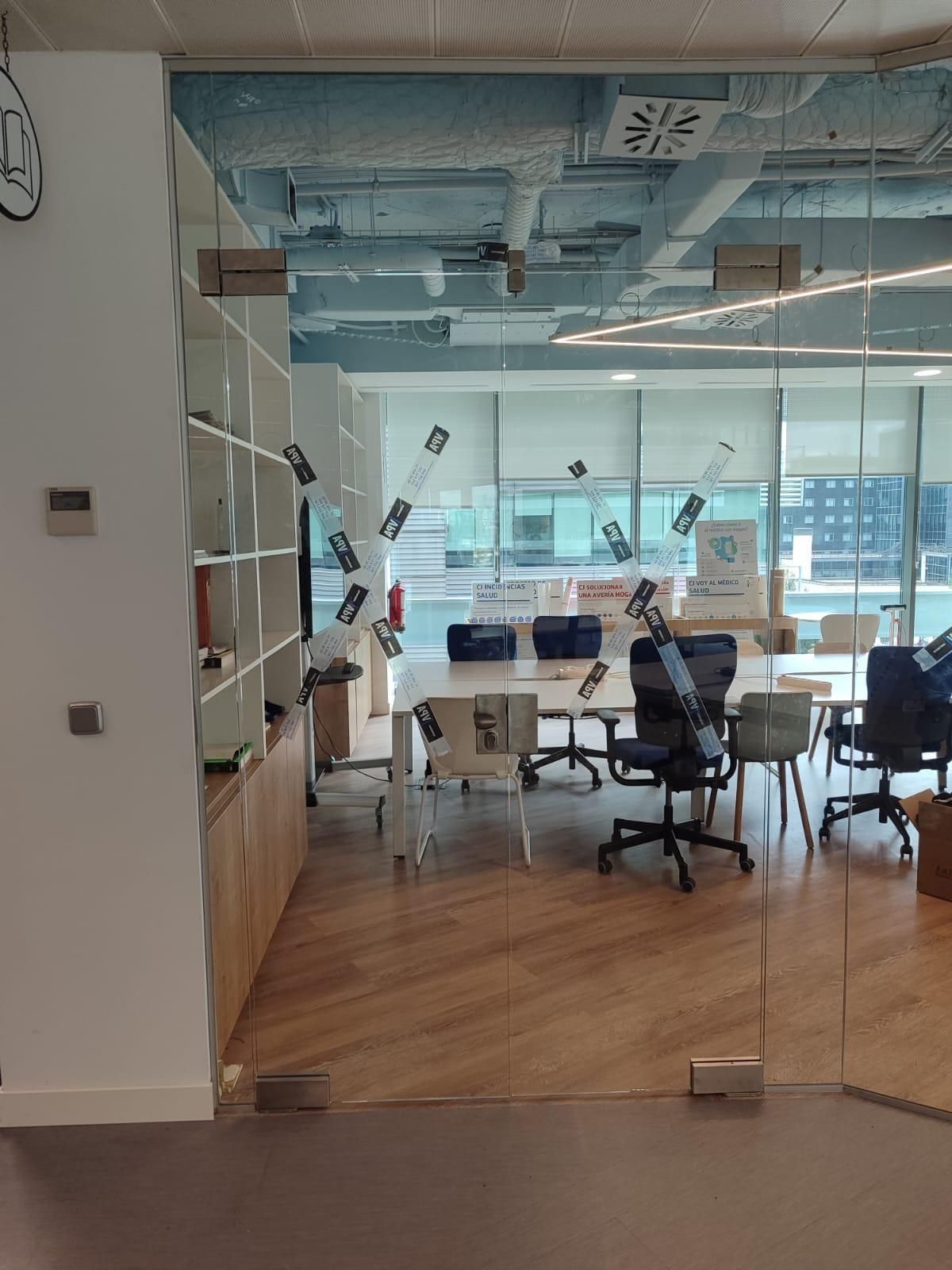 Oficina con paredes de cristal, escritorios, sillas y vistas a la ciudad.