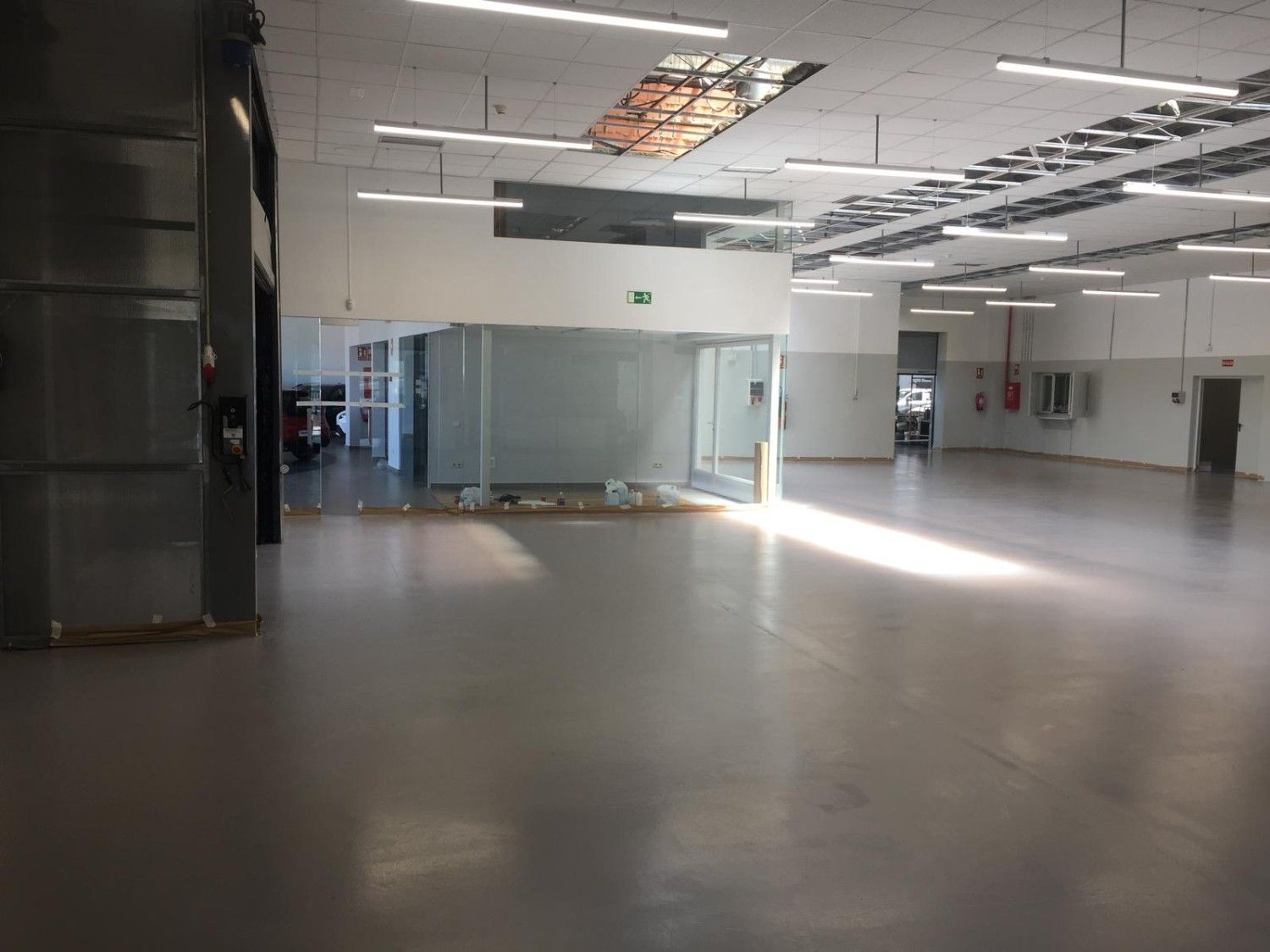 Local comercial interior vacío con suelo gris, tabiques de cristal y luces de techo a la vista.