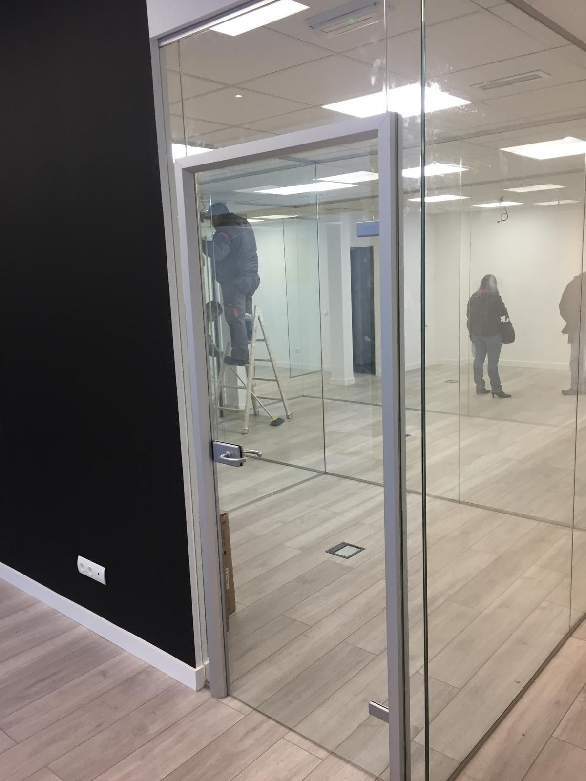 Oficina vacía con paredes de cristal, dos personas visibles y una subida a una escalera cerca de las luces del techo.