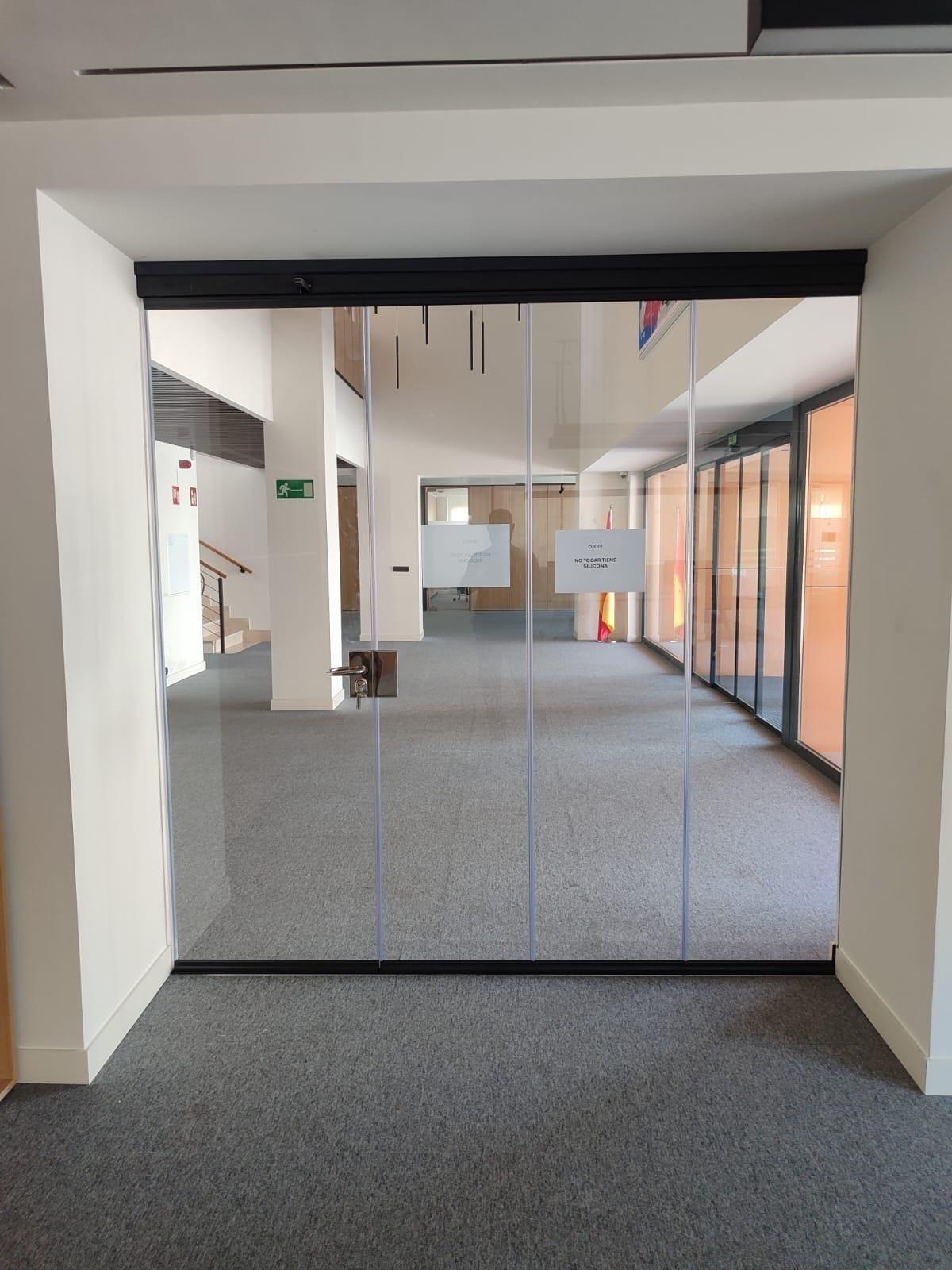 Pasillo de oficina vacío con puertas de cristal y moqueta gris, visto a través de una puerta.