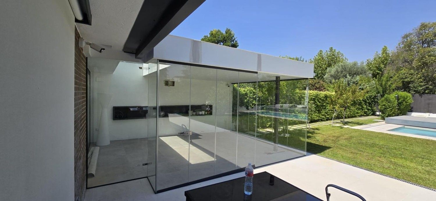 Patio moderno con barandilla de cristal, voladizo blanco y un jardín verde junto a una piscina.