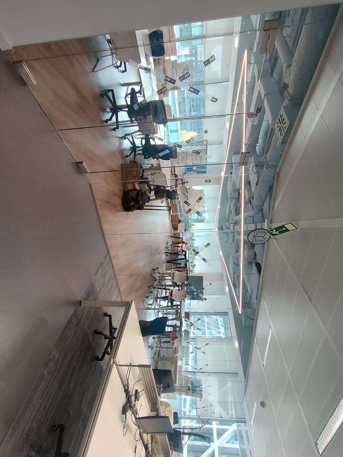 Interior de oficina moderno y concurrido, con escritorios, sillas y paredes de cristal bajo luces de techo brillantes.