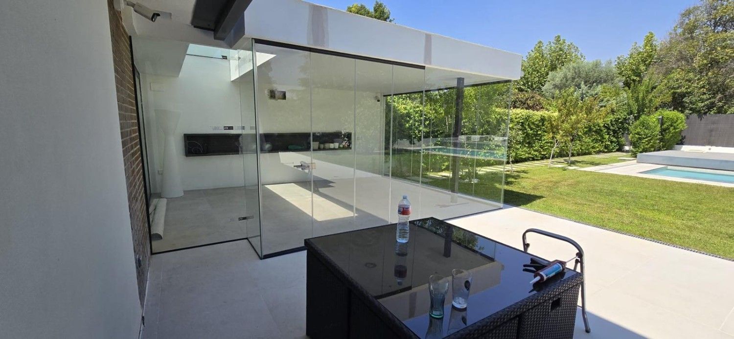 Patio moderno con pared de cristal, mesa y sillas negras, y un jardín trasero con césped.