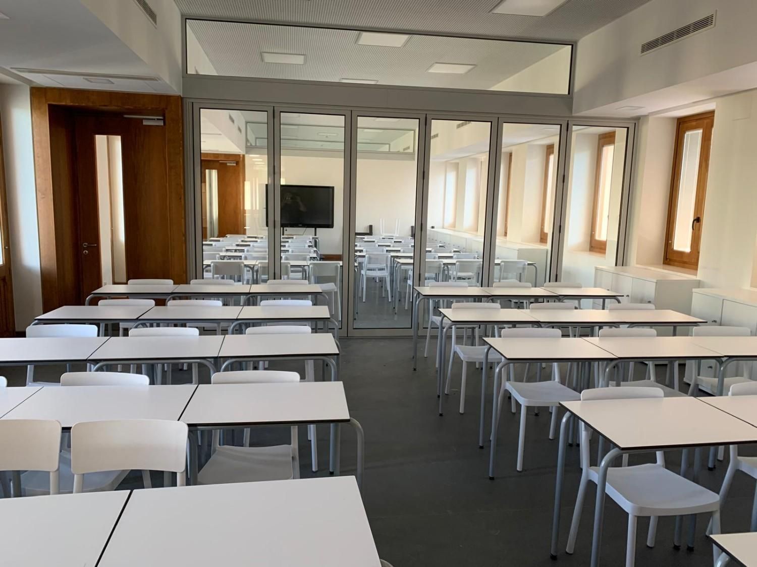 Aula vacía con filas de pupitres y sillas blancas, grandes ventanales y una mampara de cristal al fondo.