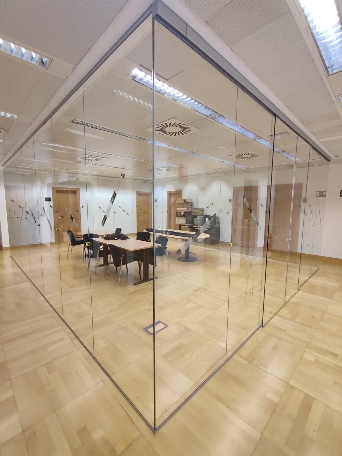 Cabina de oficina con paredes de cristal, escritorios y sillas en una habitación luminosa con suelo de madera.