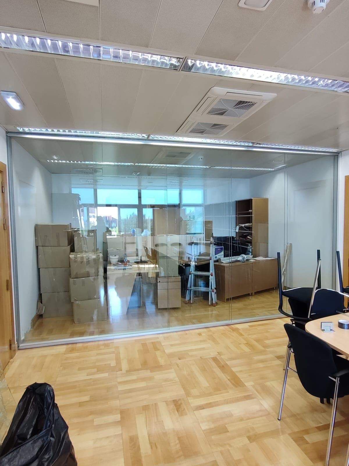 Interior de oficina con pared de cristal, cajas apiladas, escritorios y sillas sobre un suelo de madera.
