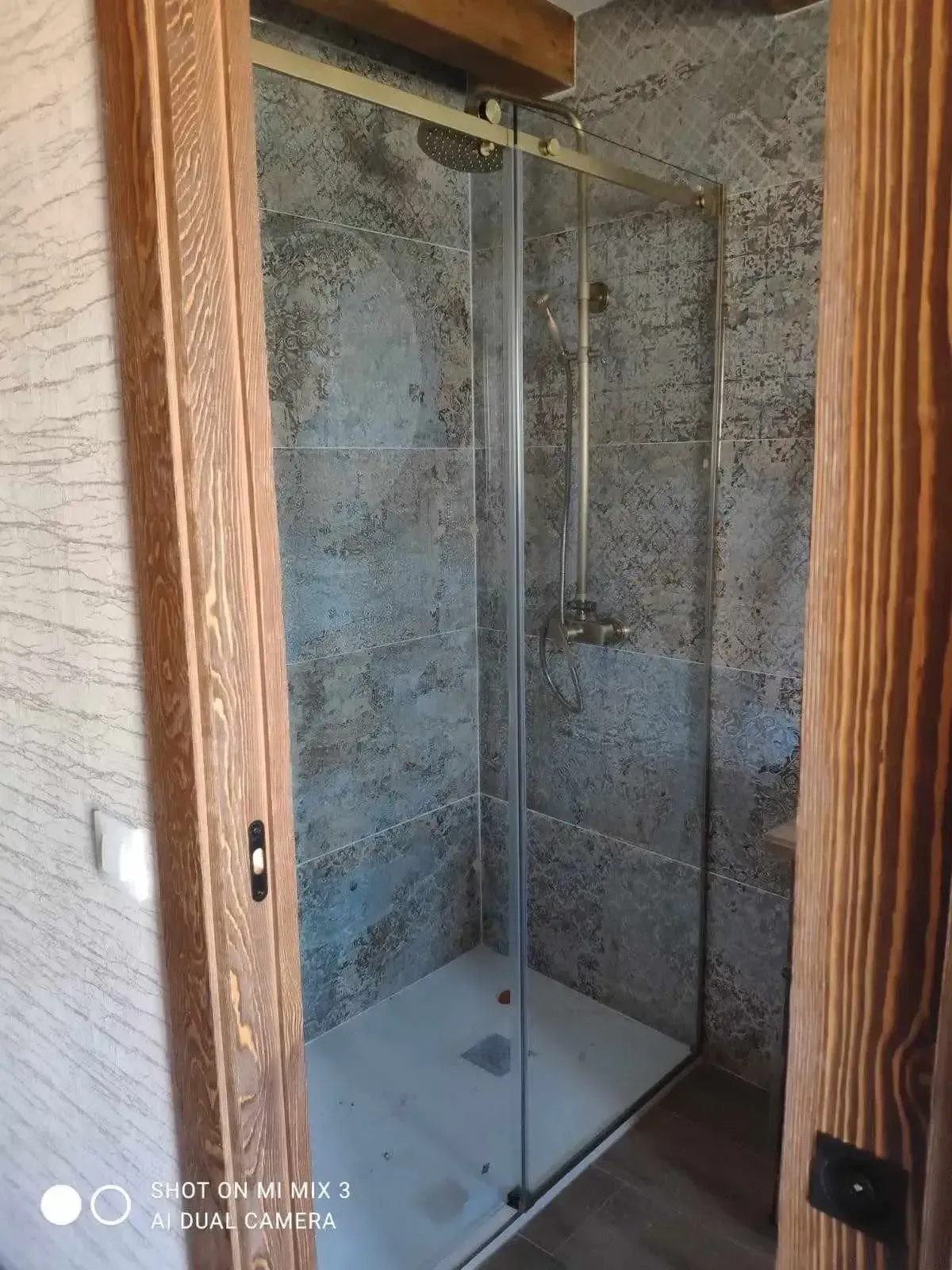 Cabina de ducha de cristal con azulejos blancos y paredes de piedra gris enmarcadas en un marco de puerta de madera.