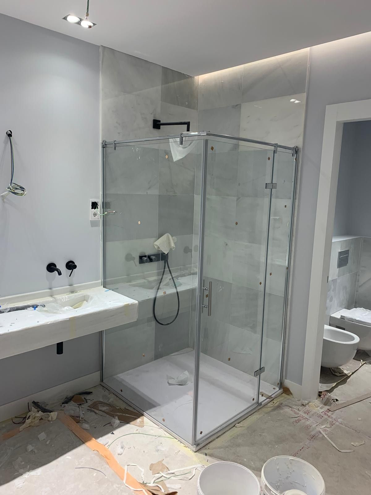 Baño moderno con cabina de ducha de cristal, lavabo mural, inodoro y baldosas de suelo sin acabado.