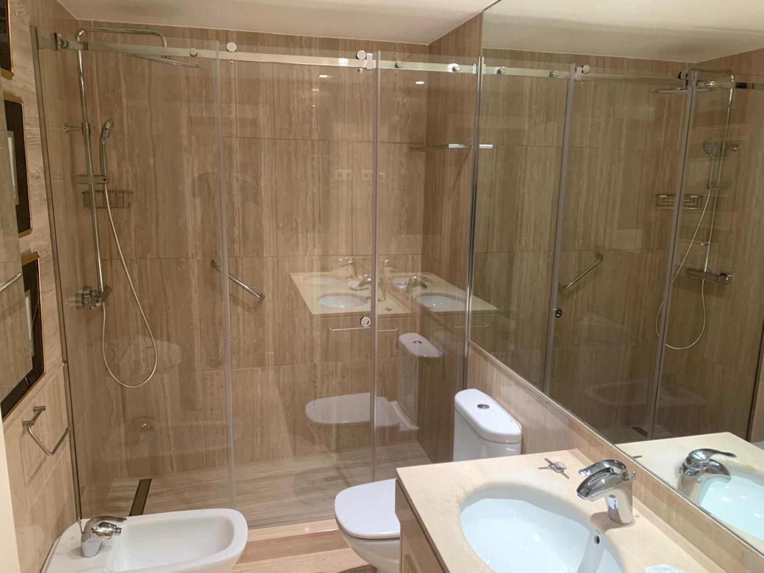 Baño moderno con ducha de cristal, paredes de azulejos beige, inodoro, bidé y tocador con doble lavabo.