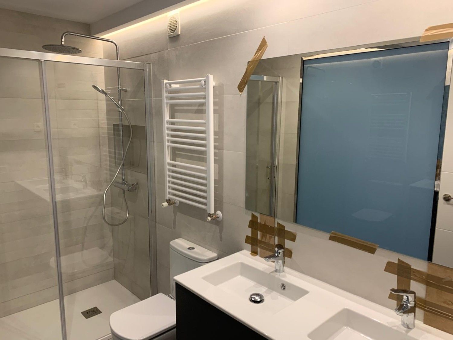 Baño moderno con ducha de cristal, radiador toallero blanco, lavabo, inodoro y cortina de ventana azul esmerilada.