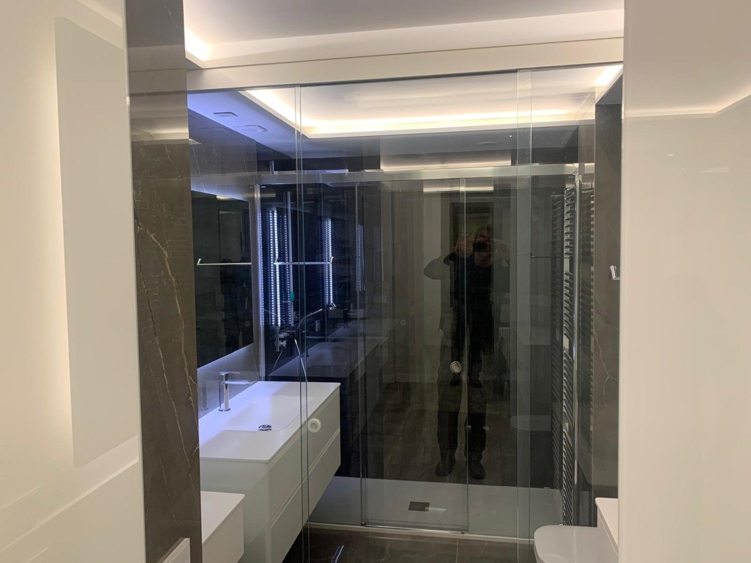 Baño moderno con cabina de ducha de cristal, lavabo blanco y pared con espejo que refleja la habitación.