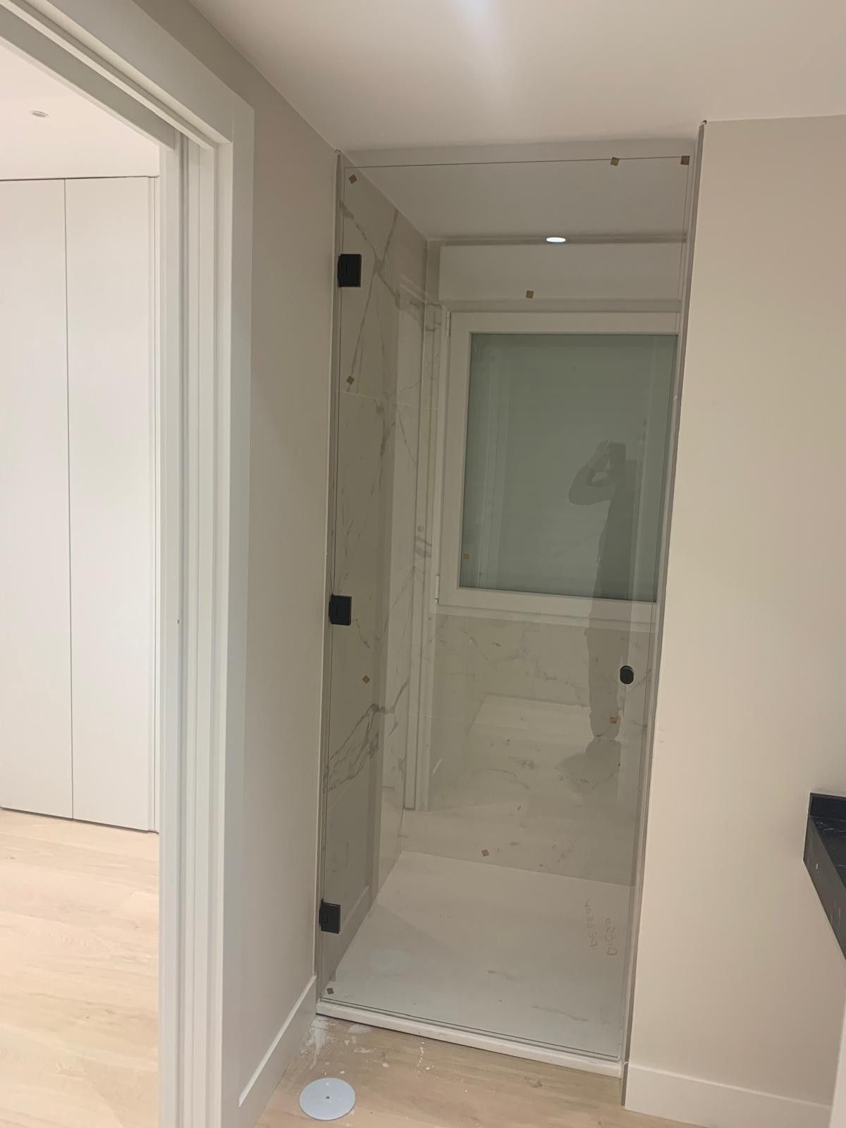 Baño con cabina de ducha de cristal y azulejos blancos, visto desde un pasillo.