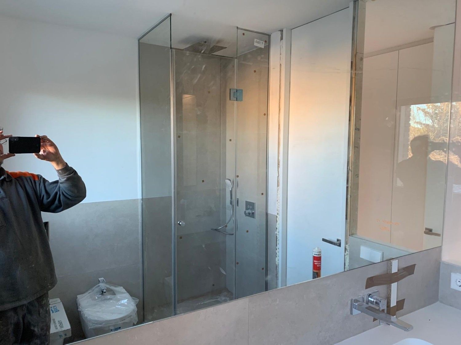 Baño con cabina de ducha de cristal, espejo de pared y lavabo; una persona está tomando una foto a la izquierda.