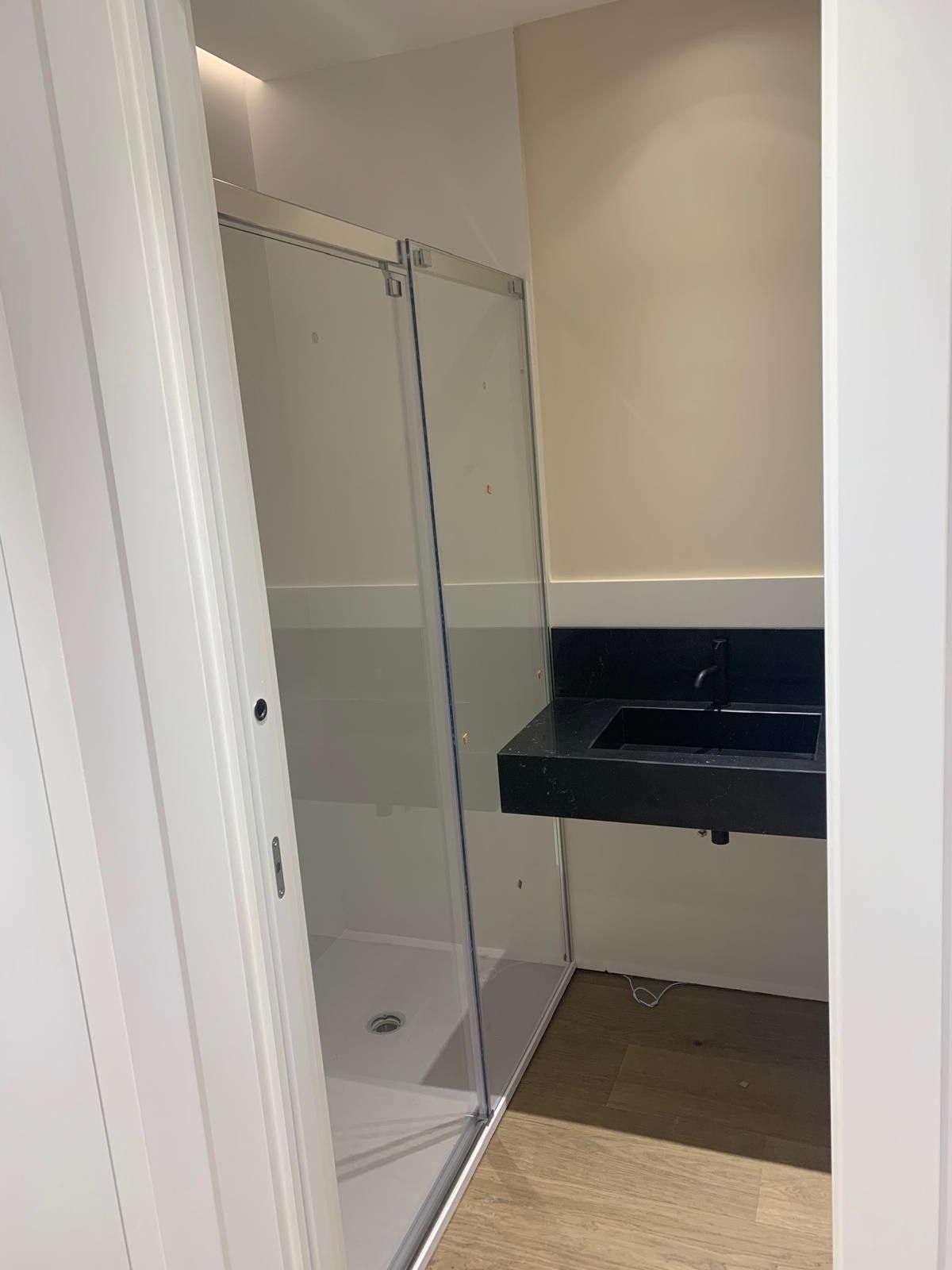 Baño pequeño con ducha de cristal, lavabo negro montado en la pared y suelo de baldosas color beige.