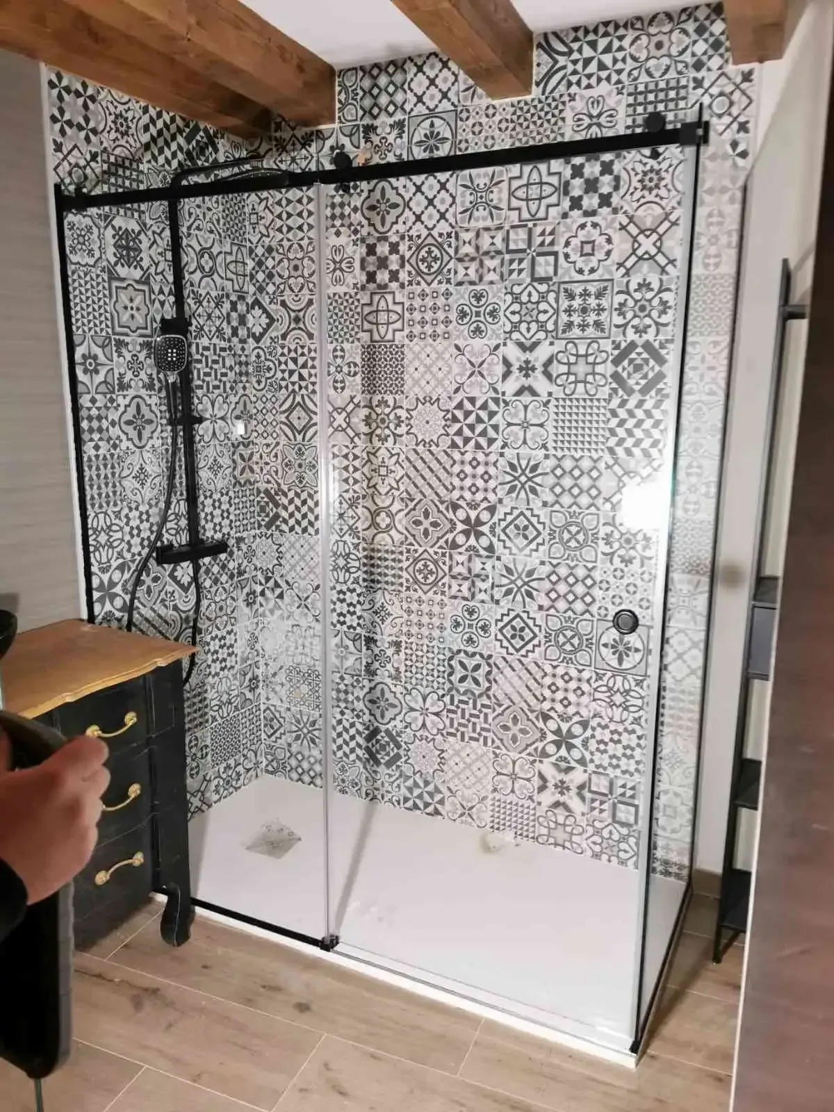 Mampara de ducha de cristal en un baño con paredes de azulejos estampados y vigas de madera en el techo.