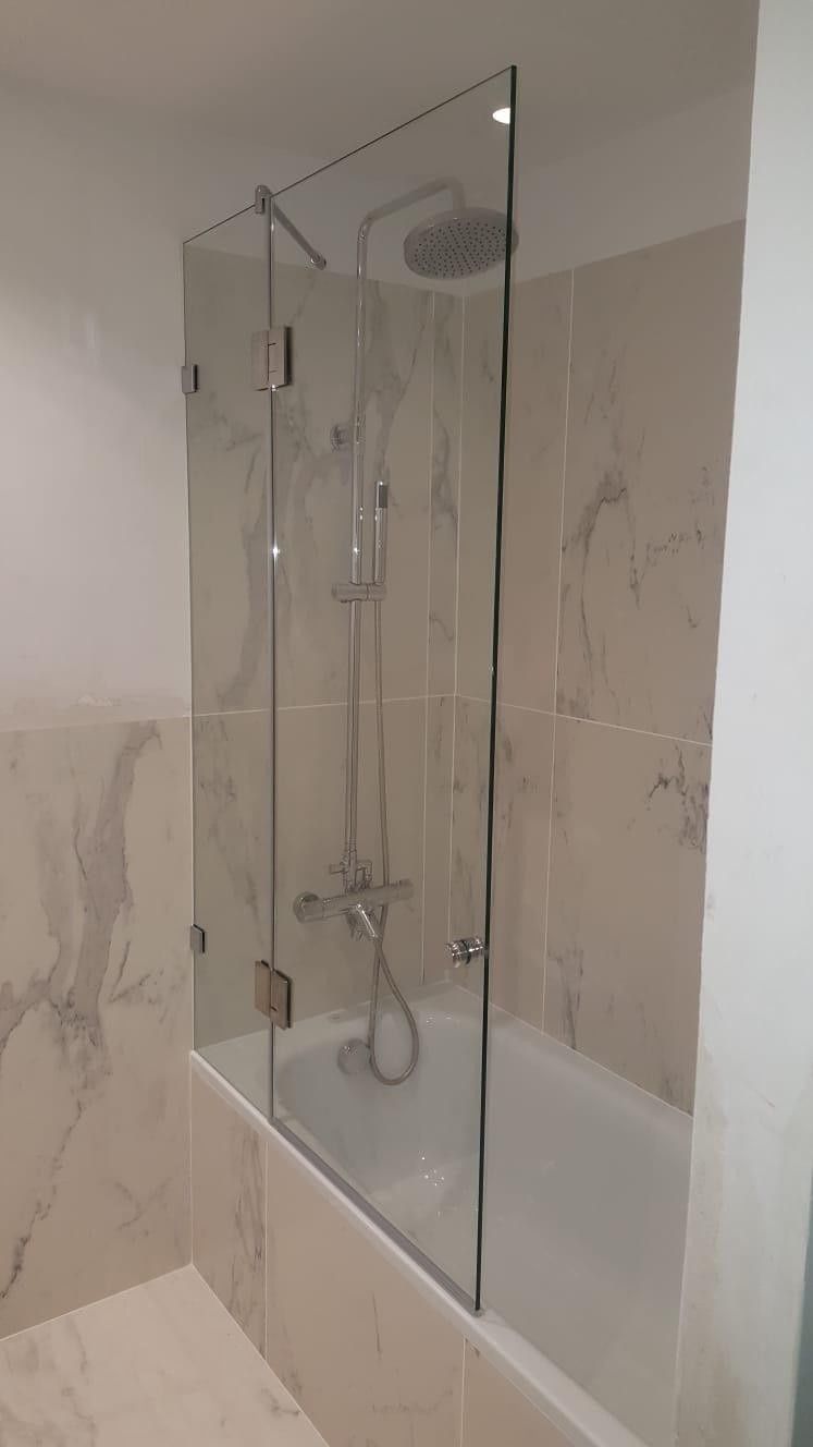 Mampara de ducha de cristal sobre una bañera blanca en un baño alicatado, con cabezal de ducha mural.