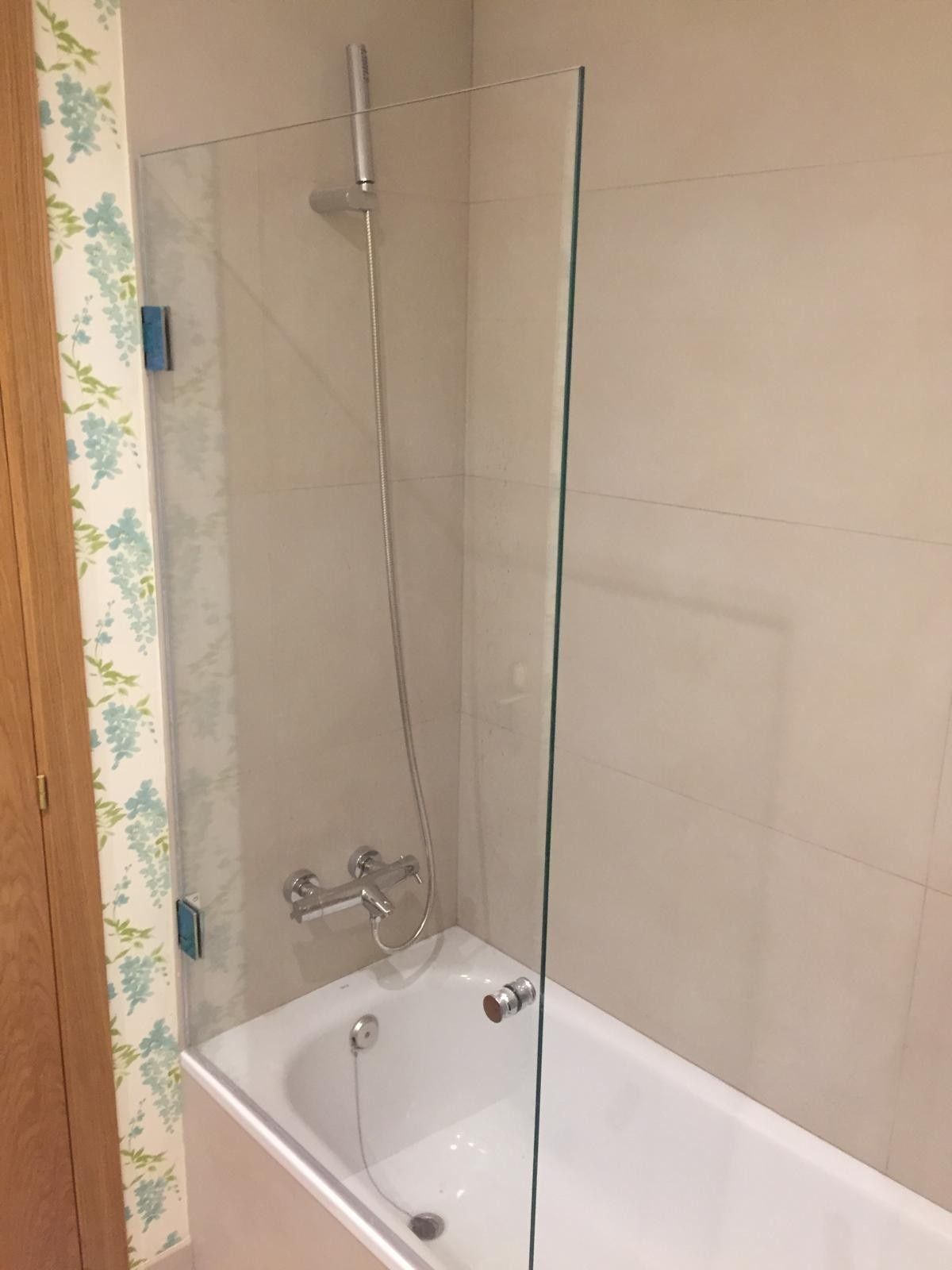 Ducha de baño sobre una bañera blanca con mampara de cristal y paredes alicatadas en color beige.