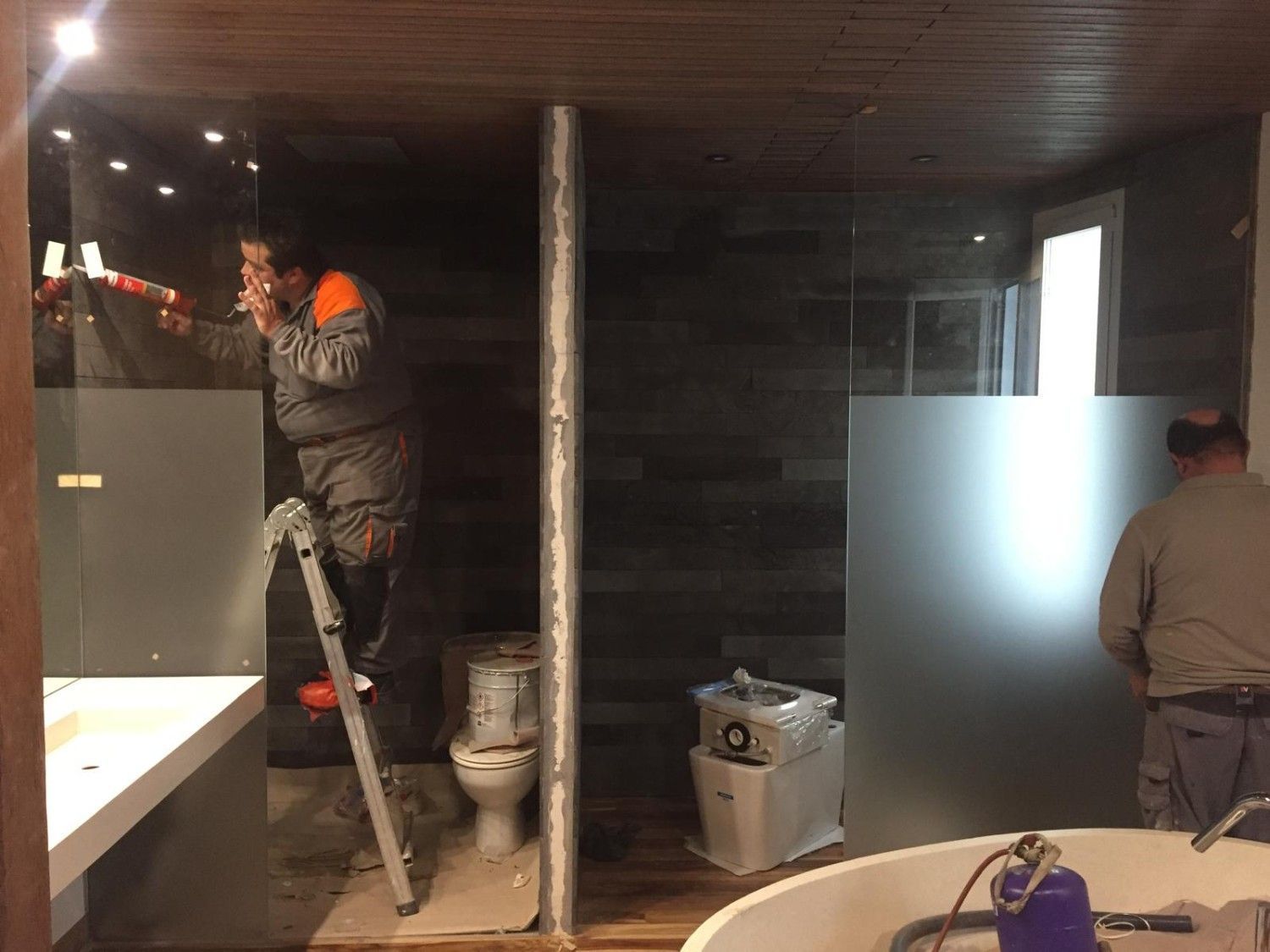 Dos trabajadores instalan accesorios en un baño con poca luz que está siendo renovado, utilizando escaleras, herramientas