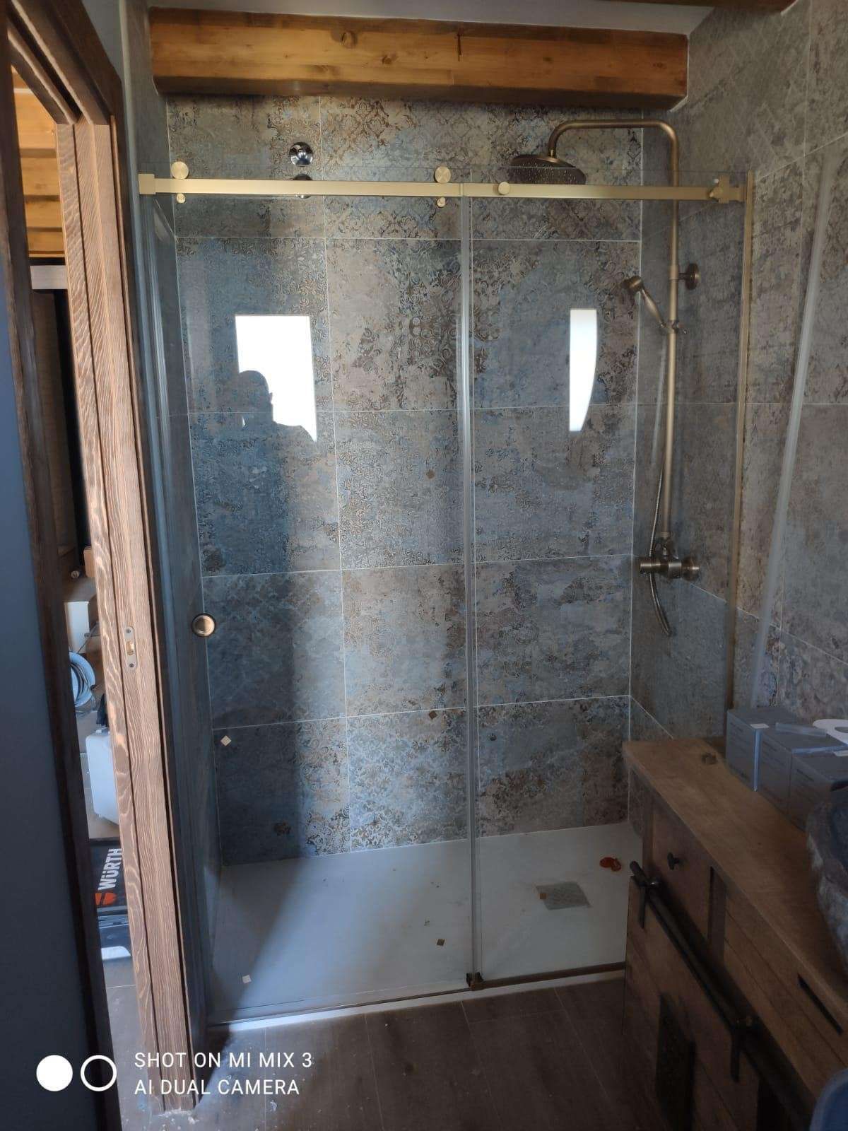 Cabina de ducha de cristal en un baño rústico, con paredes de azulejos moteados y una viga de madera en el techo.