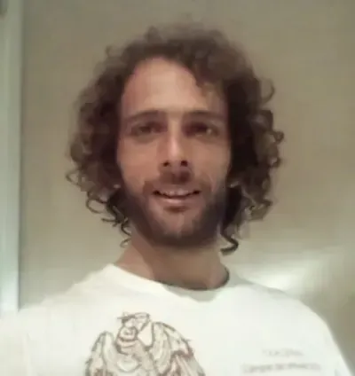 Una persona de pelo rizado con una camisa blanca sonríe en el interior de una vivienda, con una pared lisa de fondo.