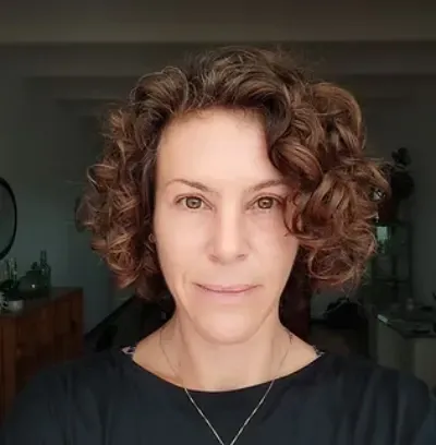 Primer plano de una persona con cabello castaño rizado, vestida con una blusa negra y un collar, mirando a la cámara.