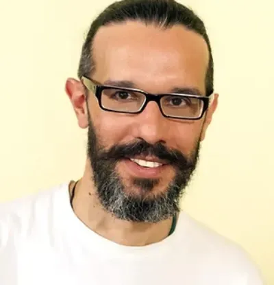 Hombre sonriente con gafas y barba, vestido con una camisa blanca, sobre un fondo claro.